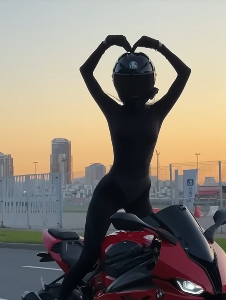 Törölték Moto Tanya Tiktok fiókját. A képen Moto Tanya menet közben áll a motoron és elengedett kormánnyal egy nagy szivet formál a kezével.