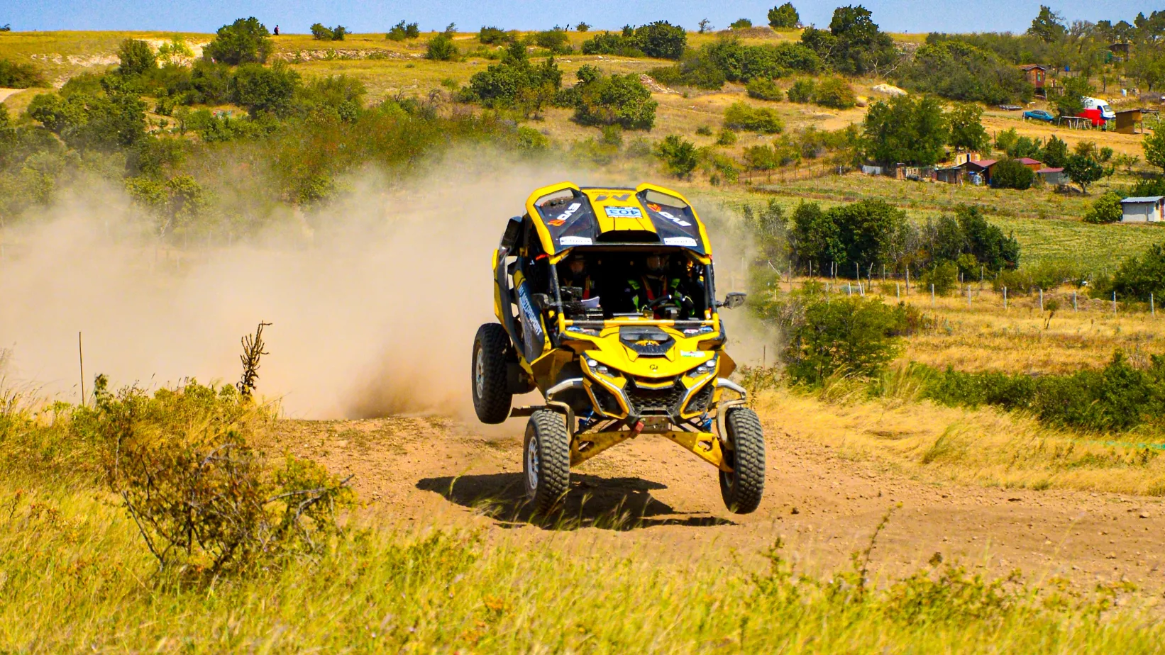 FIA Európa-kupa keretében megrendezett HunGarian Baja Rally Várpalota mellett a versenypályán a Tési dombi ugratón ugrat egy versenyautó ami épp a levegőben van, nagy por van mögötte. Akciódús fénykép amin a versenyautó színe citromsárga és fekete.