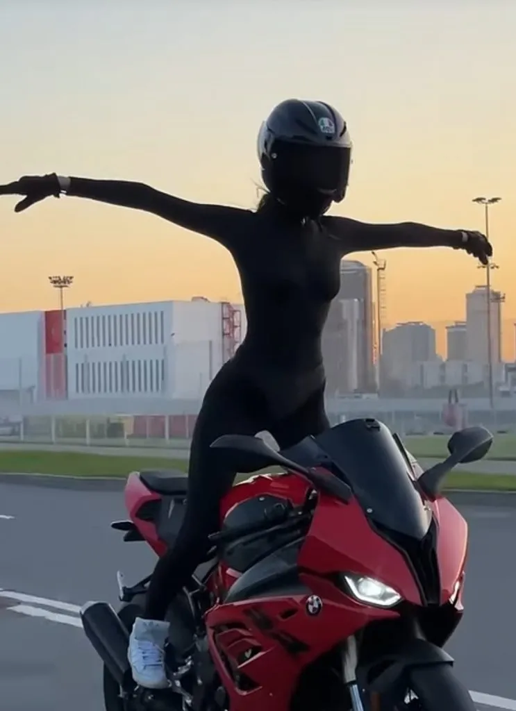 Moto Tanya menet közben áll a piros BMW S1000RR motorján elengedett kormánnyal és szét tett kezekkel fekete ruhában.