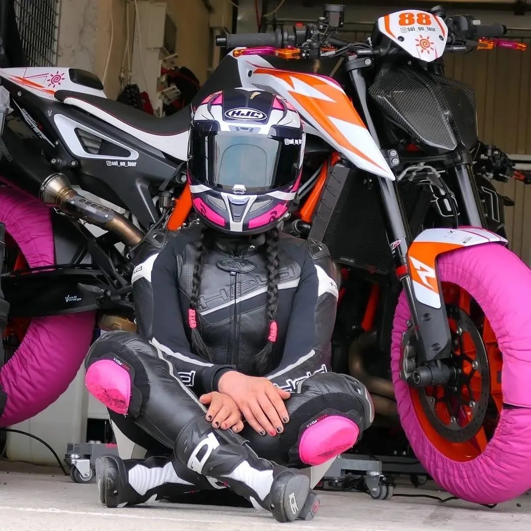 Egy KTM Duke motor előtt ül motoros ruhában egy hölgy nekitámaszkodva a motornak. A hölgy törökülésben és bukósisakban ül. A KTM motorkerékpáron rózsaszínű gumimelegitők vannak felrakva.